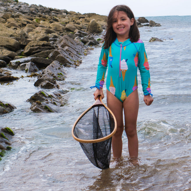 A girl in an UV Protective Long Sleeve Swimsuit Green Ice Cream is standing in the sea with a fishing net, smiling at the camera.